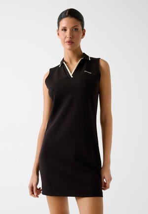 Kobieta w bezrękawną czarną sukienkę polo z białym obszyciem i logo „GUESS”, stojąca na jednolitym jasnym tle.