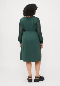 Grønn kjole med lange, gjennomsiktige ermer som har sikksakk-mønstre. Stoffet har en teksturert finish og er smalnet inn i midjen med et belte. Svarte platåhæler.