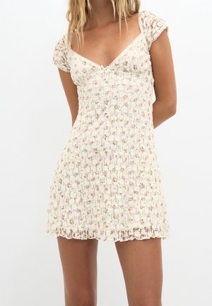 Femme portant une robe courte blanche en dentelle florale avec des manches courtes et un col en V, debout devant un fond blanc uni.