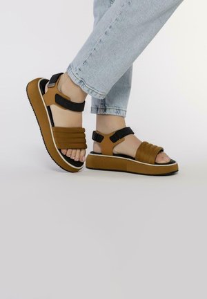 Sandales à lanières brunes et noires avec une semelle épaisse, présentant des lanières larges et des fermetures ajustables à la cheville, portées avec un jean bleu clair.