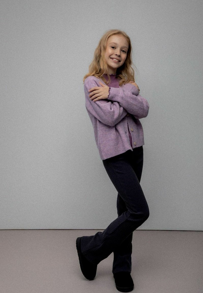 Liliowy sweter na guziki, z dzianiny, noszony na fioletowej koszuli, zestawiony z ciemnymi sztruksowymi spodniami i czarnymi butami, o fakturowanym wykończeniu.