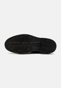 Suela negra de zapato con patrón geométrico de la banda de rodadura y nombre de la marca en relieve cerca del arco, fotografiada sobre un fondo liso y claro.