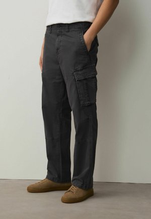 Person trägt dunkelgraue Cargo-Hose mit mehreren Taschen und braune Freizeitschuhe, steht vor einer schlichten hellen Wand.