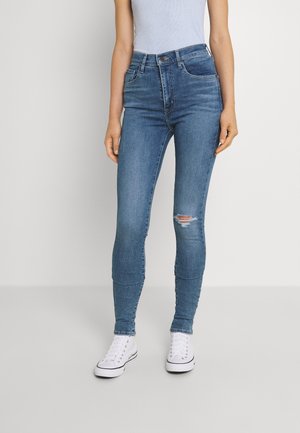 Personne portant un jean slim bleu avec une petite déchirure au genou droit et des baskets montantes blanches sur un fond uni.