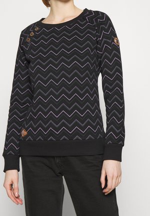 Femme portant un pull noir à manches longues avec des motifs en zigzag violet et trois boutons sur l'épaule, assorti à un pantalon noir.