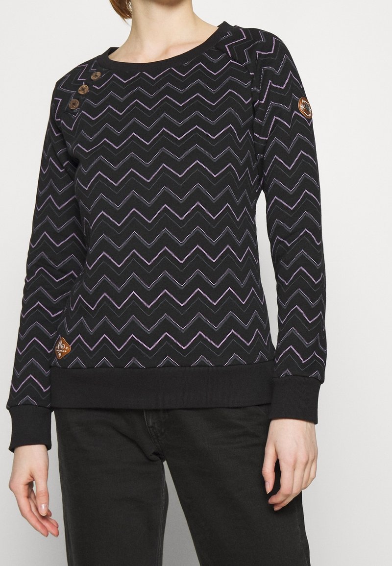 Femme portant un pull noir à manches longues avec des motifs en zigzag violet et trois boutons sur l'épaule, assorti à un pantalon noir.