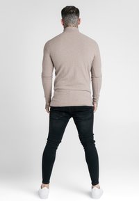Man med hals-tatuering iklädd beige ribbad polotröja, svarta skinny jeans och vita sneakers, stående vänd bort från kameran på en neutral bakgrund.
