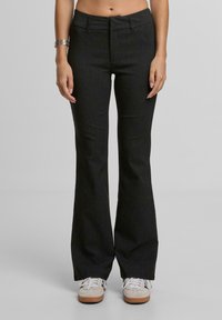 Žena v černých pruhovaných rozšířených kalhotách a bílých teniskách, stojící s uvolněnýma rukama podél těla na jednobarevném pozadí.