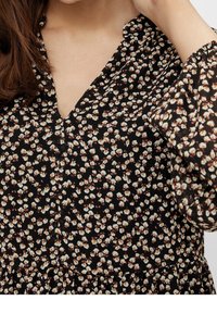 Blouse noire à fleurs avec col en V, manches longues bouffantes, et petits motifs floraux blancs et roses sur un tissu léger. Fini texturé.