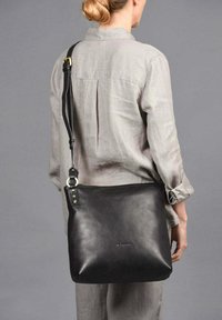 Bolso de hombro de cuero negro con una textura suave, que cuenta con una correa con acentos en dorado y herrajes de botón, exhibido sobre un fondo gris.