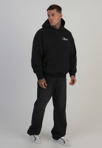 Felpa nera con cappuccio, tasca frontale e logo bianco, abbinata a jeans scuri e sneakers bianche, con vestibilità morbida.