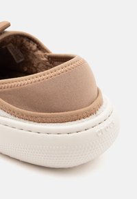 Brauner Slipper aus weichem Textilgewebe, mit Fleecefutter und weißer strukturierten Sohle, die mit dem eingeprägten Markennamen "Crocs" versehen ist.