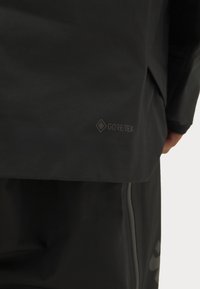Czarna kurtka GORE-TEX o gładkim, matowym wykończeniu i subtelnym logo przy dolnym brzegu. Projekt jest minimalistyczny, z prostym krojem.