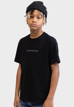 Ung person iført en sort beanie og en sort t-shirt med teksten "REDEFINED REBEL", stående foran en enkel hvid baggrund.