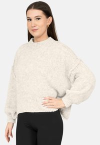 Vrouw die een lichtgrijze grove gebreide trui met ballonmouwen en zwarte leggings draagt, poseert met één hand op haar heup.