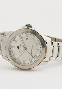 Silver Tommy Hilfiger automatic wristwatch with metal band, white dial, date display showing 22, and luminescent hour markers and hands.