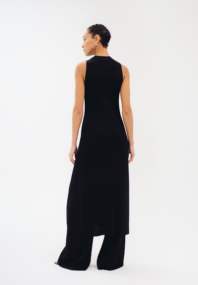 Schwarzes, geripptes Strickkleid mit hohem Kragen, ärmellosem Design und seitlichen Schlitzen, kombiniert mit weit geschnittenen schwarzen Hosen für eine schlanke Silhouette.
