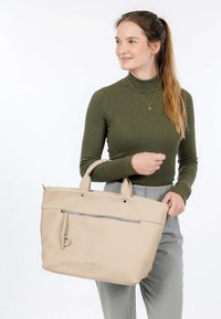 Sac fourre-tout en cuir beige avec une finition texturée, poignées doubles et une poche zippée à l'avant. Le modèle porte un haut côtelé vert olive.