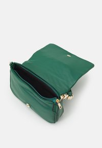 Zielona skórzana torebka na ramię z założonym klapem, złotymi akcentami, zamknięciem na zamek błyskawiczny wewnątrz i gładką teksturą. Wnętrze torebki jest czarne.