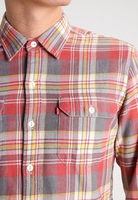 Camicia a quadri rossa e grigia con design a bottoni, dotata di due tasche sul petto e di un piccolo tag rosso con logo sulla tasca sinistra.