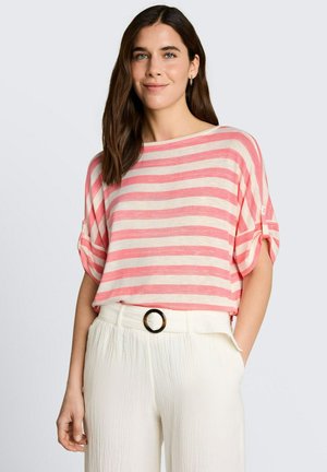 Kobieta z długimi brązowymi włosami, ubrana w różowo-biały paskowany krótki rękaw i białe spodnie z wysokim stanem oraz paskiem z okrągłą klamrą.