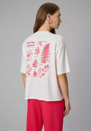 Kobieta w białej koszulce z czerwonymi botanicznymi nadrukami i napisem „Feel The Earth 4F” na plecach, zestawiona z jaskrawo różowymi spodniami.