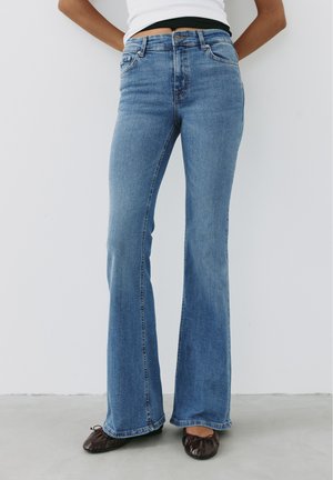 Femme portant un jean flare taille haute bleu clair avec un top blanc, debout contre un mur gris clair, portant des chaussures plates marron foncé.