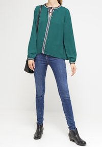 Kvinna som bär en grön långärmad blus med mönstrad trim, smala blå jeans, svarta ankelstövlar och bär en svart axelväska.