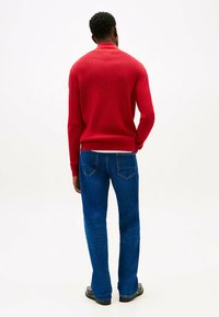 Pull en tricot rouge avec un col montant, associé à un jean bleu évasé. La texture semble côtelée avec des détails de bords et d'ourlet contrastants.