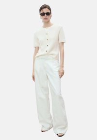 Knit, cream-colored short-sleeve top with front buttons, paired with wide-leg white pants, worn with black sunglasses.