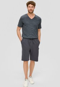 T-shirt gris foncé en v, en tissu texturé, associé à un short à cordon assorti. Le modèle porte des baskets blanches, les mains dans les poches.