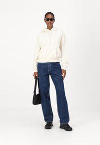 Cream hoodie with kangaroo pocket, paired with loose blue jeans and black sneakers; accessorized with black sunglasses and a shoulder bag.