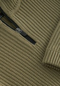 Kaki zip-up tröja med strukturerad stickad tyg, ribbade ärmmuddar och en svart dragkedja. Designen har diagonala och vertikala mönster.