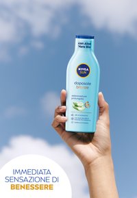 Bottiglia di latte dopo sole NIVEA Sun, plastica blu, forma ovale, testo bianco e arancione, con grafica di aloe vera, contro un cielo nuvoloso.