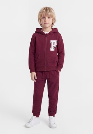 Enfant blond debout portant un sweat à capuche bordeaux zippé avec un F blanc et un pantalon de jogging assorti avec des baskets blanches.