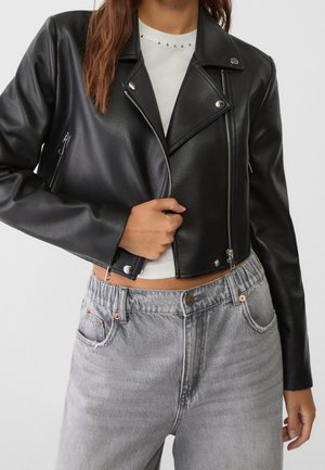 Femme portant une veste en cuir noire courte sur un haut blanc et un jean taille haute gris clair, ajustant la fermeture éclair de la veste.