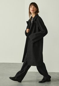 Woman walking indoors wearing a long black textured coat, striped shirt, wide-leg black pants, and black leather shoes against a plain background.
