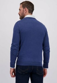 Blauwe gebreide trui met lange mouwen, geribbelde manchetten en zoom. Witte blouse met kraag met stippen zichtbaar bij de halslijn en manchetten.