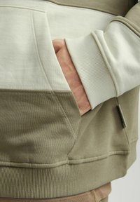 Sweatshirt beige et vert olive avec une manchette côtelée, une poche kangourou et des textures de tissu contrastantes. Détails de couture notables.