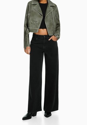 Vrouw draagt een korte bikerjack in olijfgroen, een zwart kort topje, zwarte wijde high-waist jeans en zwarte laarzen met hak.