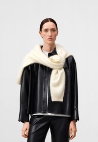 Schwarze Lederjacke in cropped Länge über einem weißen Pullover mit einem übergroßen cremefarbenen Schal. Glatte Textur, auffällige Knöpfe und entspannte Passform.
