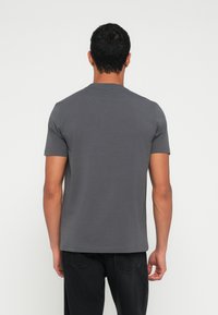 Άνδρας που στέκεται με την πλάτη, φοράει απλό σκουρόχρωμο γκρι κοντομάνικο μπλουζάκι και μαύρα τζιν παντελόνια πάνω σε λευκό φόντο.