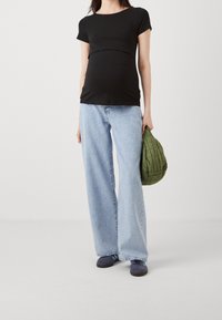 Zwarte zwangerschapsblouse met een ronde hals en korte mouwen, gecombineerd met lichtblauwe broek met wijde pijpen. Houdt een groene gewatteerde tas vast.