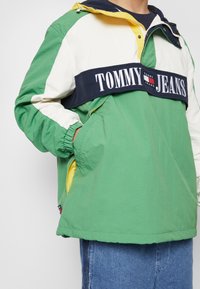 Vēja slānis ar zaļiem, baltiem un tumši zilajiem krāsu bloku, elastīgām piedurknēm, kapuci un izteiktu "TOMMY JEANS" logotipu priekšpusē.