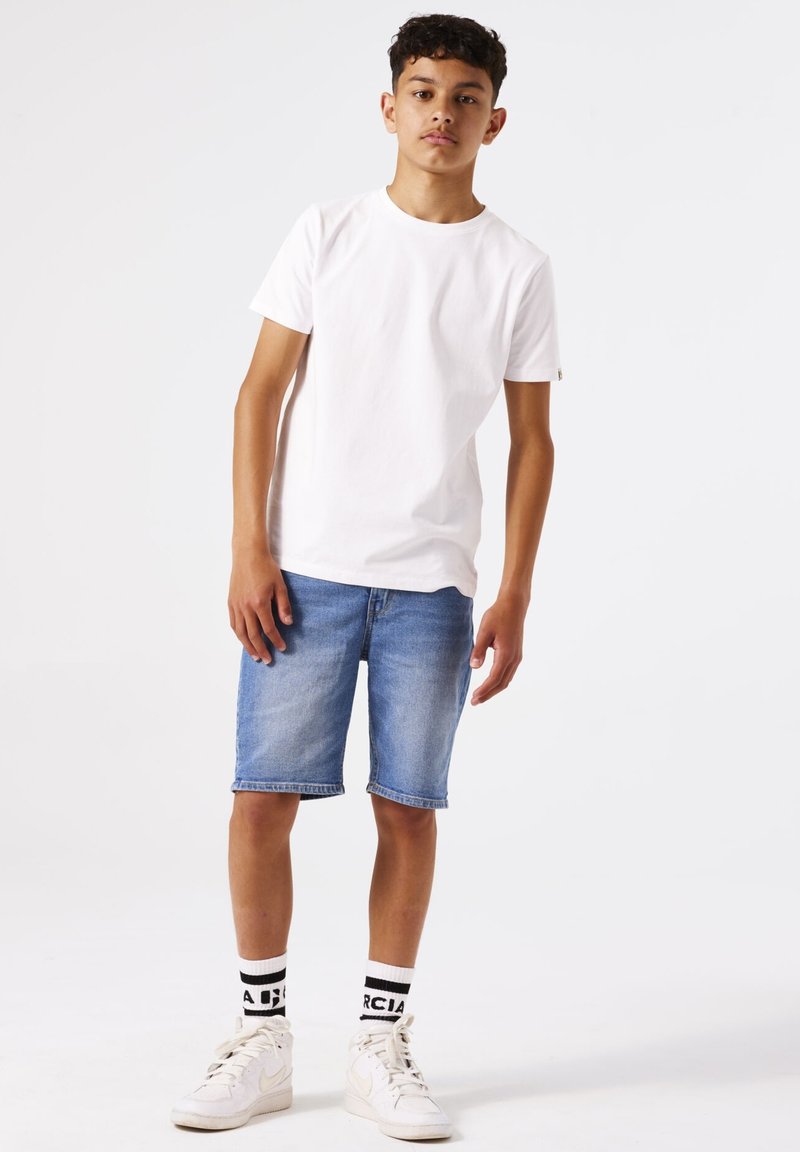 Camiseta blanca de algodón, pantalones cortos de mezclilla azules, zapatillas altas blancas y calcetines de caña corta en blanco y negro con texto. Un atuendo simple y casual.