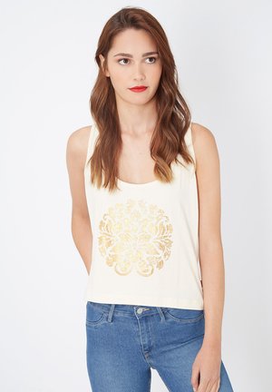Camiseta sin mangas color crema con un estampado floral dorado en el centro. Presenta un escote redondo y un corte holgado. Combinada con unos jeans azules.