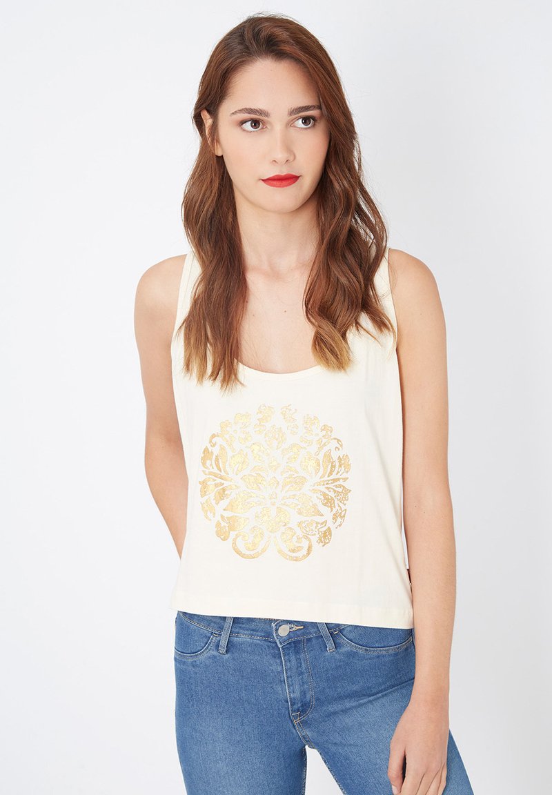 Camiseta sin mangas color crema con un estampado floral dorado en el centro. Presenta un escote redondo y un corte holgado. Combinada con unos jeans azules.