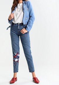 Giacca in suede azzurro chiaro, camicia bianca con bottoni, jeans in denim a vita alta con ricami floreali rossi e blu, e scarpe rosse senza lacci.