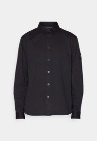 Camisa de manga larga de algodón negra con cuello abotonado, cierre frontal de botones y un parche en la manga izquierda.