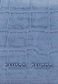 Gaiši zils ādas izstrādājums ar krokodila rakstu un gludu tekstūru, uz kura pacelts "SAMSOE" zīmola nosaukums līdzīgā tonī, kas uzlabo dizainu.